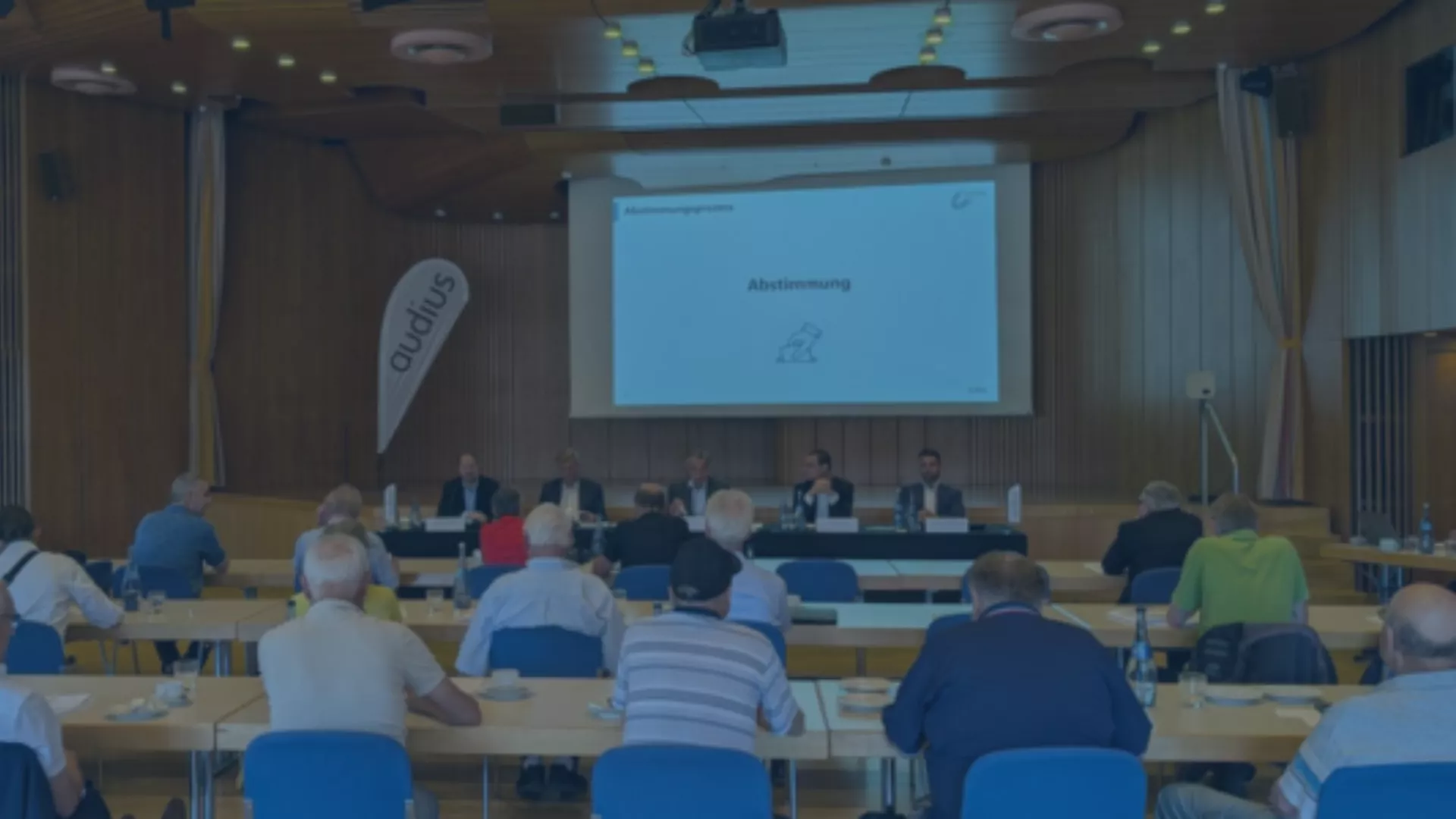 Teilnehmer einer Hauptversammlung sitzen vor einer Bühne mit Podium und Leinwand mit dem Titel „Abstimmung“
