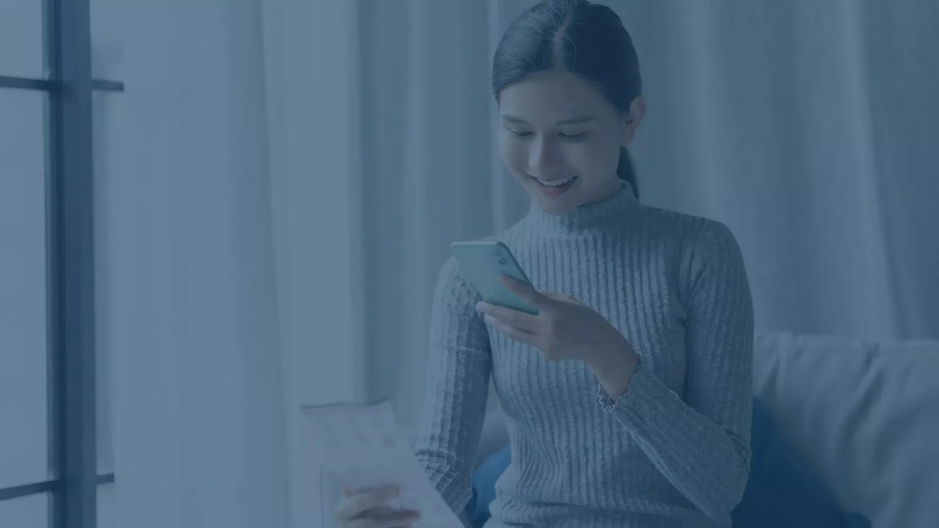 Frau sitzt auf dem Sofa, hält ein Dokument in der Hand und nutzt ihr Smartphone, um Informationen abzurufen oder zu prüfen.