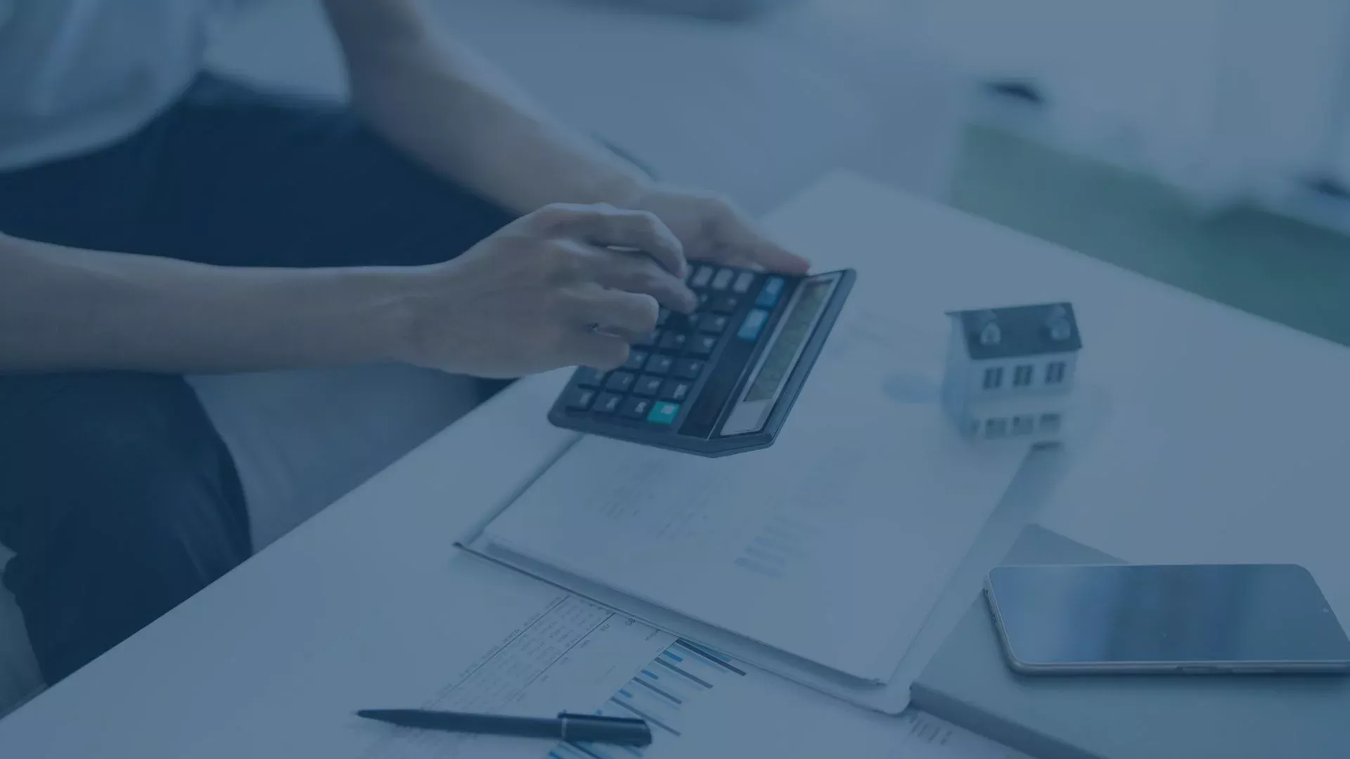 Person berechnet finanzielle oder steuerliche Daten mit einem Taschenrechner, daneben liegen Dokumente und ein kleines Modellhaus.