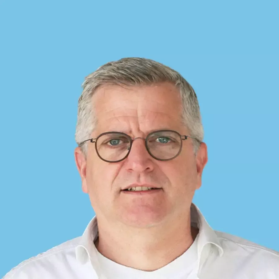 Porträt eines Mannes mit Brille und grauen Haaren vor hellblauem Hintergrund.